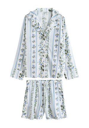 Hosszú barna hajú nő, fehér virágmintás pizsama szettben, kék és zöld függőleges mintákkal, beltéren, áttetsző függönyök és növények közelében áll.