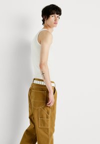 Top in cotone bianco a coste, pantaloni utility marroni con tasche multiple e una cintura bianca con fori neri. Modello che guarda a destra, mostrando il profilo e i dettagli dell'outfit.
