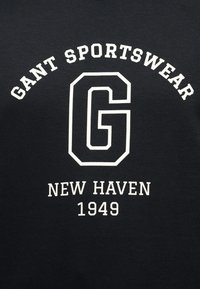 Μαύρο ύφασμα με λευκό κείμενο: «GANT SPORTSWEAR» κυρτό πάνω από ένα μεγάλο περιγραμμικό «G» και «NEW HAVEN 1949» από κάτω.
