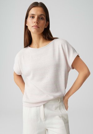 Frau mit langem braunem Haar, trägt ein hellrosa kurzärmeliges Oberteil und weiße hoch taillierte Hosen, steht mit den Händen in den Hüften.