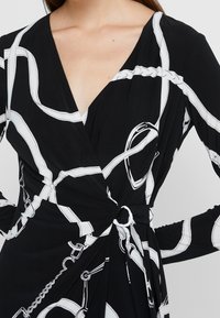 Svart wrapklänning med vit kedjemönster, långa ärmar och vriden knutdetalj vid midjan. Slät tygtextur.