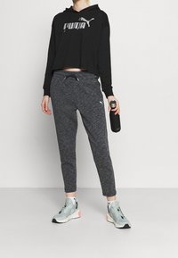 Osoba ubrana w czarną bluzę z kapturem Puma, szare spodnie dresowe Puma, miętowe sneakersy, trzymająca czarną butelkę na wodę, stojąca na tle białym.