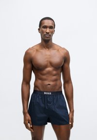 Temno modre kopalne hlače z elastičnim pasom in belim napisom "BOSS". Lahka tkanina, dolžina nad koleni in detajl z gumbom.