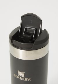 Gobelet de voyage en acier inoxydable noir isolé avec une bande métallique argentée, doté d'un couvercle à bouton-poussoir et d'un logo d'un lion ailé.