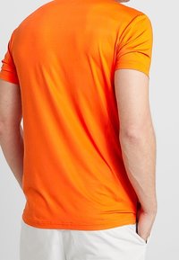 Camiseta de manga corta, que absorbe la humedad, en un vibrante color naranja, fabricada con un suave tejido sintético, con un cuello redondo y sin costuras visibles.