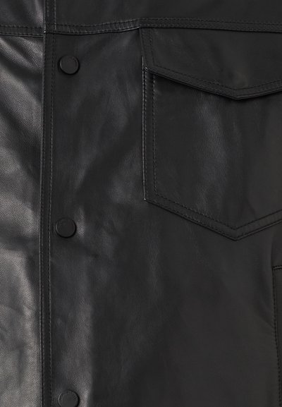 Μαύρο δερμάτινο μπουφάν με απαλό υφή, κουμπιά με πιέτα και τσέπη στο στήθος με αιχμηρό καπάκι. Καθαρές γραμμές και περιορισμένες λεπτομέρειες.
