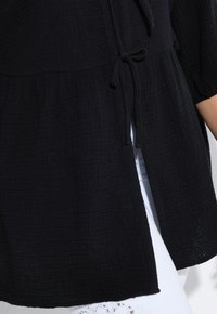 Black textured tunic with a cinched waist, featuring a drawstring tie and short sleeves. Layered design with side slits for ease of movement.