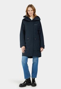 Námořnická voděodolná bunda s kapucí, na zip a rovného střihu. Nositá přes světle modré džíny a černé boty. Hladký povrch, minimalistický design.