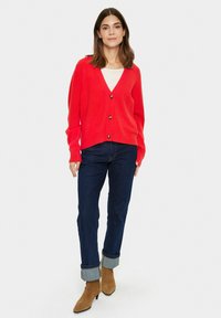 Röd kofta med V-urringning, fem knappar och långa ärmar. Kombineras med mörk jeans och beige ankelstövlar med spetsig tå.