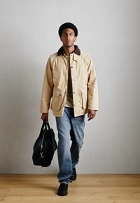 Beige jakke med brun krage, lys grønn skjorte, blå jeans, svarte sko og en svart bag. Subjekter går på tregulv.