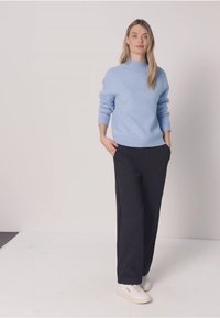 Frau steht mit den Händen in den Taschen, trägt einen hellblauen Pullover, schwarze weit geschnittene Hosen und weiße Sneakers vor einem schlichten Hintergrund.