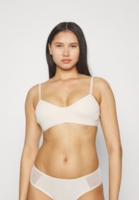 Lys beige trådløs BH med tynne justerbare stropper, glatte stoffer og V-hals-design. Modellen står mot en nøytral hvit bakgrunn.