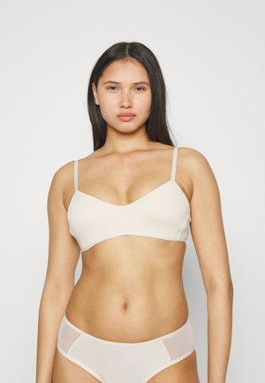 Light beige wireless bra with thin adjustable straps, smooth fabric, and a V-neck design. Model stands against a neutral white background.