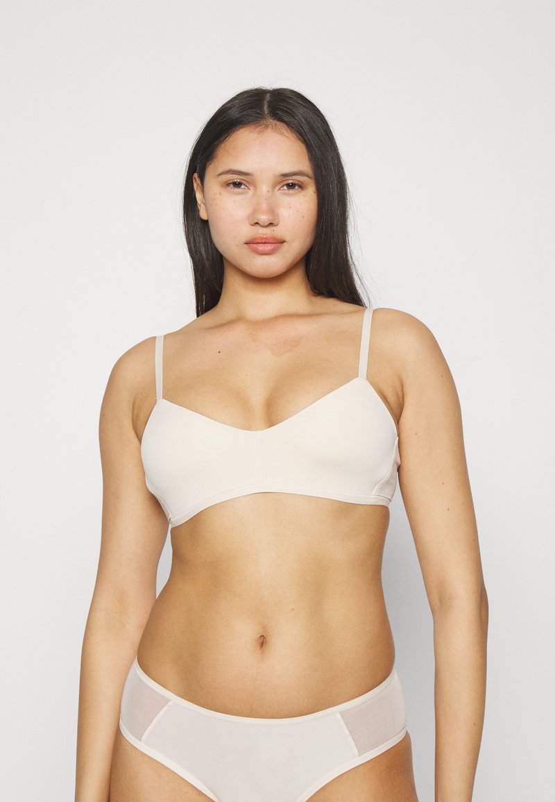 Lys beige trådløs BH med tynne justerbare stropper, glatte stoffer og V-hals-design. Modellen står mot en nøytral hvit bakgrunn.