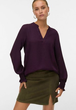 Vrouw in een paarse blouse met lange mouwen en gerimpelde manchetten, gecombineerd met een olijfgroene corduroy rok met een voorkeurspleet, poserend met haar handen bij haar heupen.