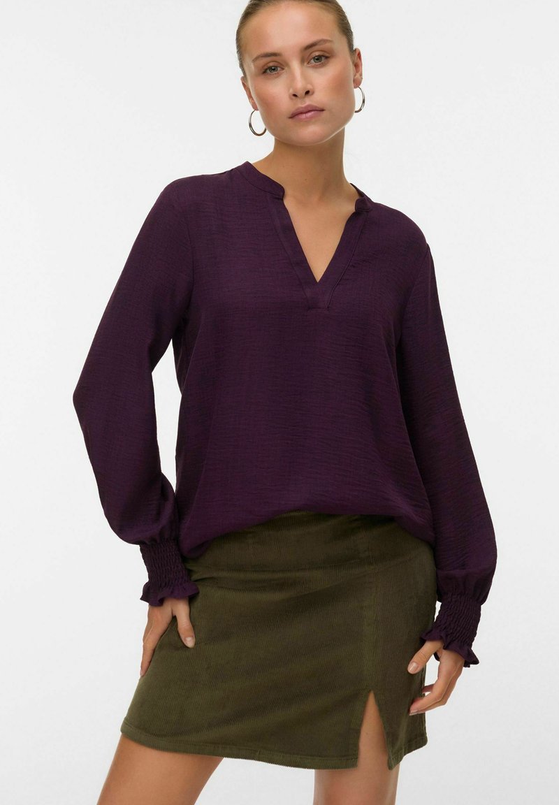 Donna che indossa una blusa lunga viola con polsini arricciati e una gonna in velluto a coste verde oliva con uno spacco frontale, in posa con le mani sui fianchi.