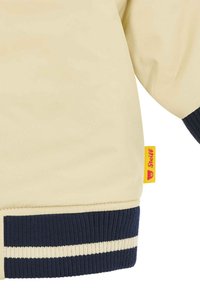Giacca bomber beige chiaro con polsini e orlo a coste blu navy. Presenta un'etichetta gialla con logo rosso cucita di lato. Materiale morbido, design sportivo.
