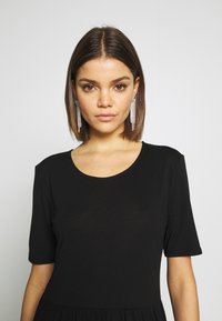 Svart kortermet kjole med rund hals og tettsittende bodice, som går over til en utsvingt hem. Modellen har på seg lange, glitrende øreringer.