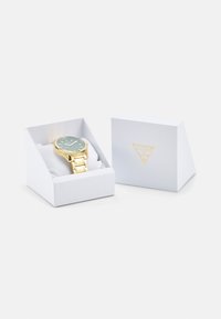 Reloj de oro con una esfera verde azulado, correa de metal y caja de exhibición blanca con acabado texturizado y logo en relieve.