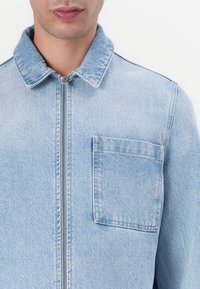 Lyseblå denimjakke med krage, glidelås foran og en lomme på venstre bryst. Har et teksturert stoff med klassisk snitt.
