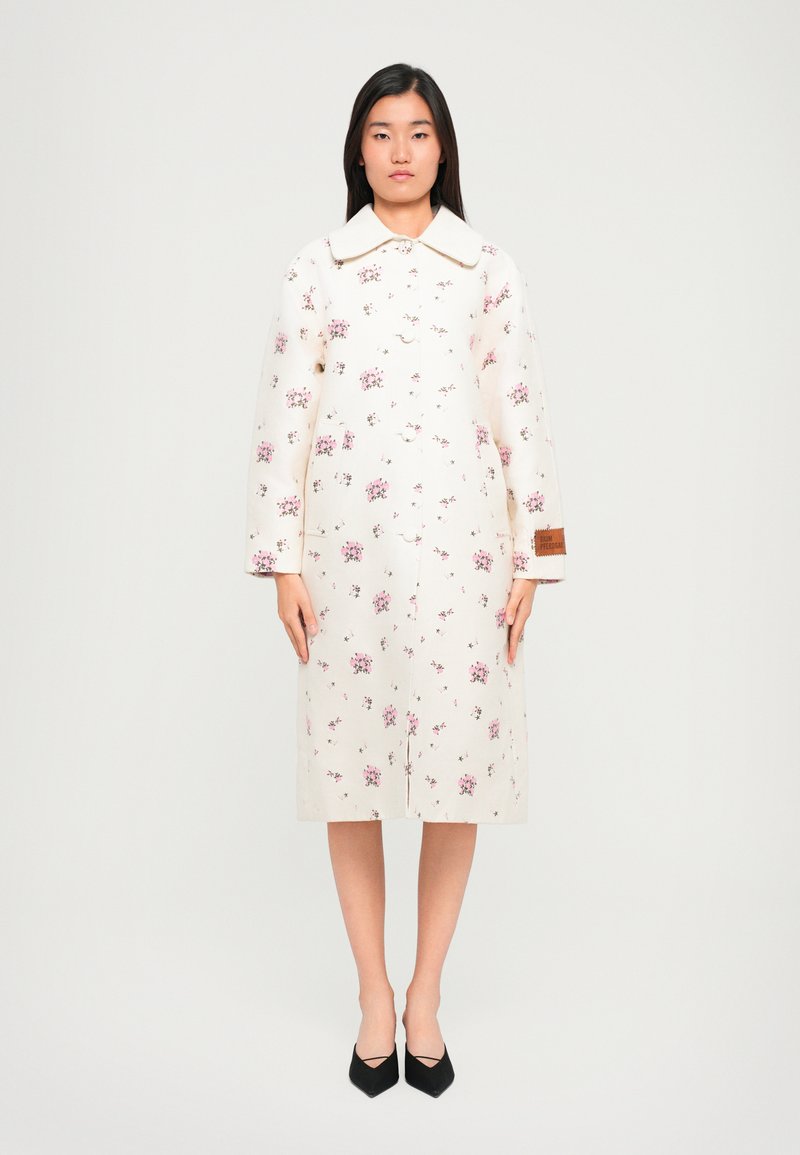 Krém színű, virágmintás kabát, galléros nyakkal és hosszú ujjal, elöl gombos záródással, a ujján egy barna színű díszcímkével.