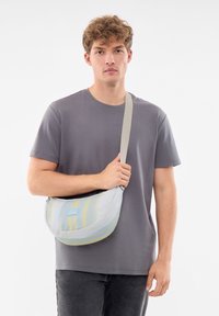 En ung man bär en grå t-shirt och svarta byxor, håller en pastellrandig axelväska med ett brett beige rem.