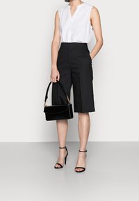 Žena v bezramínkovém bílém topu, černých šortkách s vysokým pasem po kolena, černých lodičkách na vysokém podpatku a drží černou kabelku.