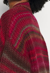 Stickad tröja i nyanser av röd, rosa och brun med ett texturerat, zicksackmönster. Har en hög, rundad halsringning och en lös passform.