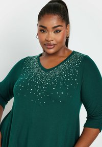 Teal V-paita, jossa on kolmannen osan hihat, koristeltu hopea- ja kultapilkuilla, jotka ovat hajautettuina rinnan ja hartioiden alueelle.