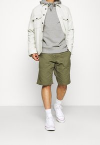 Felpa grigia chiara sotto una giacca bianca, abbinata a pantaloni corti verde oliva. Sneakers e calze bianche completano l'outfit.