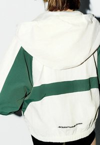 Biała i zielona kurtka przeciwdeszczowa z kapturem, z wyrazistym poziomym wzorem pasków, elastycznymi mankietami i siateczkową podszewką. Logo na plecach.