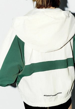 Biała i zielona kurtka przeciwdeszczowa z kapturem, z wyrazistym poziomym wzorem pasków, elastycznymi mankietami i siateczkową podszewką. Logo na plecach.