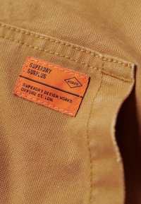 Tela color beige con una etiqueta naranja cosida que dice "Superdry Surplus" y "Superdry Design Works, Oxford St, LDN." El texto es negro en la etiqueta.