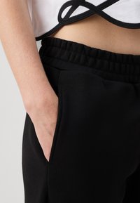 Juodi sportiniai kelnės su elastinga liemene ir šonine kišene, derinami su trumpu baltu viršumi, kuriame yra juoda apdailos juosta.