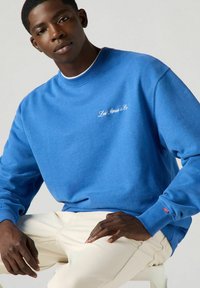 Sudadera azul hecha de una mezcla de algodón, con cuello y puños acanalados, logo bordado en blanco en el pecho y una etiqueta roja en la manga.