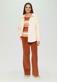 Lichtroze button-up jas over een gestreepte trui in meerdere kleuren. Bruine corduroy broek en beige platform sneakers maken de outfit af.