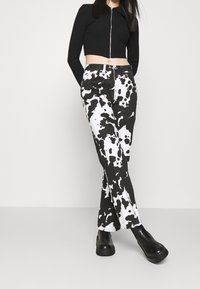Černý zip-up cropped top a kalhoty s potiskem krávy v černé a bílé barvě. Kalhoty mají kapsy a rovný střih. Modelka má na sobě masivní černé boty.