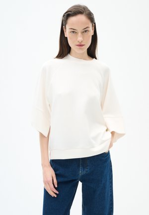Felpa oversize color avorio chiaro con maniche corte, dalla consistenza morbida e dal collo rotondo, abbinata a jeans a gamba larga blu scuro.