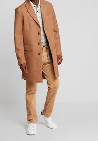 Manteau en mélange de laine marron avec revers à cran et boutons noirs, porté sur une chemise à motifs et un pantalon en velours côtelé beige, associé à des baskets blanches.