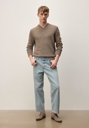 Mladý muž stojí s rukami v kapsách, oblečený v hnedom svetri s výstrihom do V, svetlomodrých džínsoch a béžových ležérnych topánkach na jednoduchom pozadí.