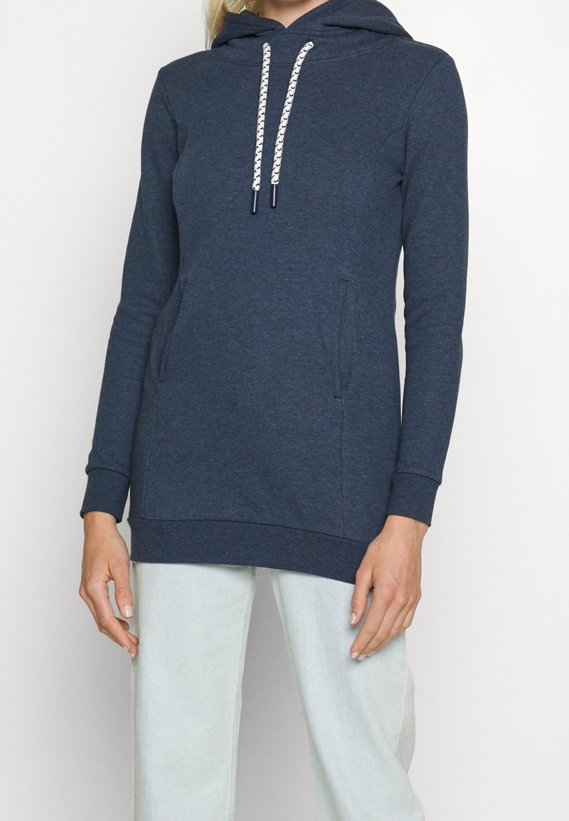 Femme portant un sweat à capuche bleu marine à manches longues avec cordons blancs et un jean bleu clair devant un fond uni.