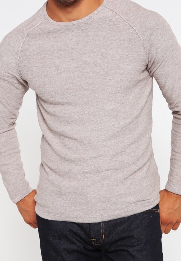 Homme portant un pull léger gris clair à manches longues avec un pantalon foncé, debout, mains près des hanches, sur un fond blanc uni.