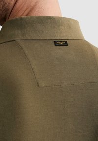 Primer plano del cuello y hombro de un hombre que lleva una camisa polo texturizada de color verde oliva con una pequeña etiqueta de logo negro y dorado que dice "American Classic".