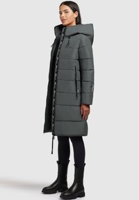 Manteau long en duvet gris foncé avec capuche, fermeture éclair et poches latérales. Présente des coutures horizontales et des accents décoratifs en bande noire.