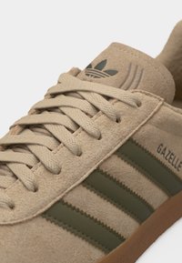 Μπεζ αθλητικό παπούτσι Adidas Gazelle με ρίγες σε ελαιδί πράσινο, μπεζ κορδόνια και σόλα από καουτσούκ, που δείχνει υφή από σουέντ υλικό και το λογότυπο της μάρκας στη γλώσσα.