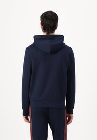 Sweatshirt azul marinho feito de tecido de algodão misto, com capuz ajustável por cordões, punhos e bainha canelados, combinado com calças de treino escuras com riscas laterais em bordô.