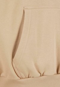 Gros plan sur un sweat à capuche beige présentant une poche kangourou et un ourlet élastique côtelé, mettant en valeur la texture du tissu et les détails de couture.