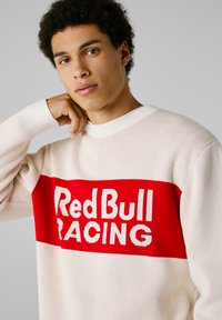 Cremfarbener Pullover mit einem auffälligen roten horizontalen Streifen und dem Schriftzug "Red Bull RACING" in Weiß. Rippkragen und -bündchen sorgen für zusätzliche Strukturen.
