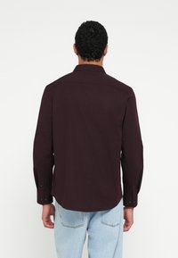 Sötét bordó, hosszú ujjú, gombos ing, hegyes gallérral és fekete gombokkal. Az anyag simának tűnik; az ing a csípőig ér.