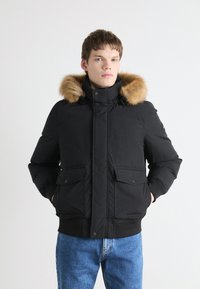 Jachetă pufos neagră cu glugă detașabilă căptușită cu blană, două buzunare frontale și manșete ribbate. Material neted și croială puțin supradimensionată.